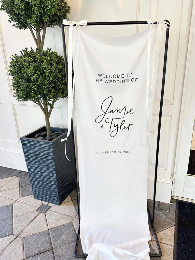 Op de afbeelding: Witte bruiloft welkom bord met zwarte tekst. Het bord staat "Welkom op de bruiloft van Jamie & Tyler, 13 september 2024".