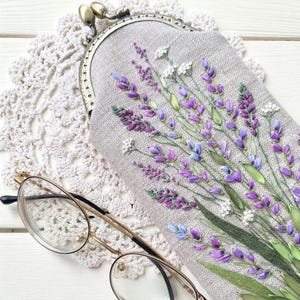 May include: A light gray glasses case with a metal clasp, featuring embroidered lavender sprigs in shades of purple, blue, and green. The case rests on a white lace doily, with gold-rimmed glasses in the foreground.