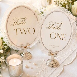 May include: Two oval table number cards with white lace trim, held by silver metal stands. One card reads Table TWO, and the other reads Table ONE with the text LEXIS & DAVID 6.13.26. The cards feature a thin brown border and elegant brown lettering.