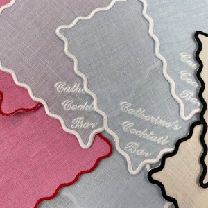 May include: Three white linen cocktail napkins with scalloped edges and white embroidery. The napkins are embroidered with the text "Catherine's Cocktail Bar".