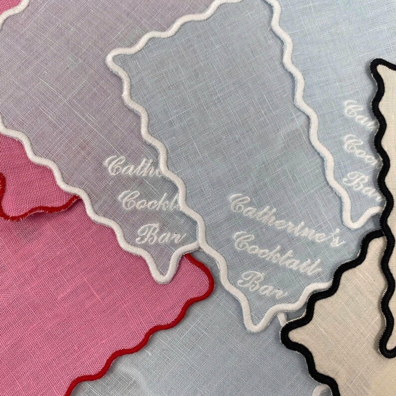 May include: Three white linen cocktail napkins with scalloped edges and white embroidery. The napkins are embroidered with the text "Catherine's Cocktail Bar".