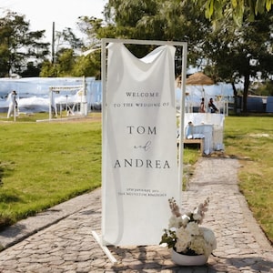 Op de afbeelding: Een wit huwelijksbord met een witte stoffen achtergrond op een wit metalen frame. Het bord staat "Welkom op de bruiloft van Tom en Andrea" met de datum "07 januari 2023" en de locatie "The Majestic Residence".