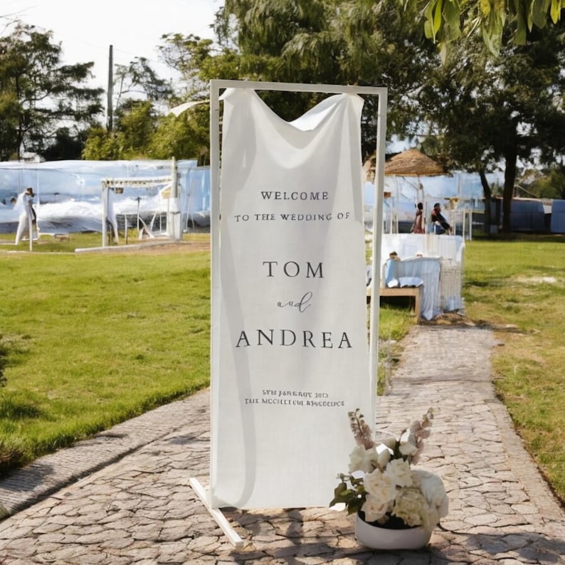 Op de afbeelding: Een wit huwelijksbord met een witte stoffen achtergrond op een wit metalen frame. Het bord staat "Welkom op de bruiloft van Tom en Andrea" met de datum "07 januari 2023" en de locatie "The Majestic Residence".