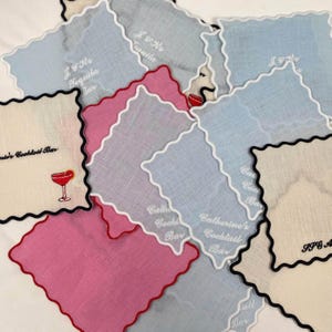 May include: A stack of light blue and cream linen cocktail napkins with scalloped edges and black or white embroidered text. The text includes "J & Jos Tequila Bar", "Catherine's Cocktail Bar", "Cath Cocktail Bar", and "all Bar".