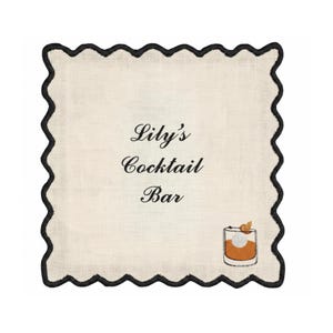 May include: A square, off-white cocktail napkin with a black scalloped border. The text "Lily's Cocktail Bar" is centered, with a graphic of a cocktail glass in the lower right corner. The napkin is made of a textured fabric.