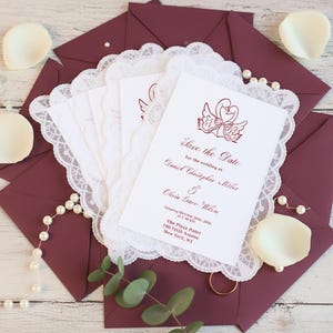 Könnte beinhalten: Weiße Save the Date-Karten mit einer Schwan-Illustration und weinrotem Text, auf einer weißen Spitzendecke. Die Karten sind von weinroten Umschlägen, Perlenschnüren und weißen Blütenblättern umgeben. Der Text lautet: Save the Date für die Hochzeit von Daniel Christopher Miller & Olivia Grace Whelen.