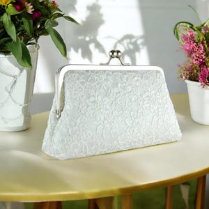 May include: A white satin clutch purse with a floral design and a silver clasp.