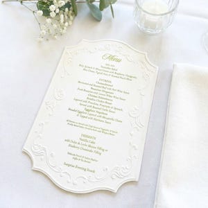 May include: A white die-cut menu card with an ornate embossed border and sage green text. The menu lists salad, entrees, and desserts including Chateaubriand, Branzino Oreganato, Chicken Saltimbocca, Eggplant Napoleon, and Vanilla Cake.