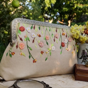 Pu&ograve; includere: Pochette beige con chiusura in metallo e ricamo floreale. I fiori ricamati sono nelle tonalit&agrave; del rosa, del rosso e del bianco, con foglie e steli verdi. La pochette ha uno stile vintage e una cornice in metallo decorativa.