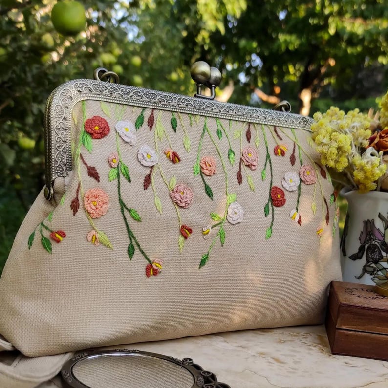 Pu&ograve; includere: Pochette beige con chiusura in metallo e ricamo floreale. I fiori ricamati sono nelle tonalit&agrave; del rosa, del rosso e del bianco, con foglie e steli verdi. La pochette ha uno stile vintage e una cornice in metallo decorativa.