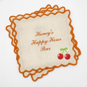 May include: Off-white cocktail napkins with orange scalloped edges. The napkins feature the text "Honey's Happy Hour Bar" in orange script, along with a pair of red cherries with green stems. A set of bar accessories.