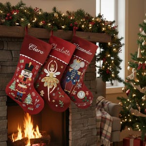 Puede incluir: Tres calcetines navideños rojos cuelgan sobre una chimenea. Cada calcetín está bordado con un diseño diferente con el tema del Cascanueces y un nombre: Clara, Charlotte y Alexander. Un árbol de Navidad y una guirnalda con luces están en el fondo.
