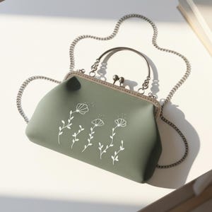 May include: A sage green handbag with a silver chain strap and a clasp closure. The handbag features a white floral design. The handle is sage green. The bag is on a white surface.