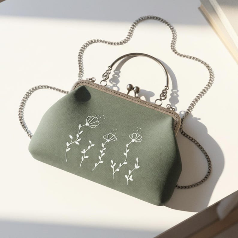 May include: A sage green handbag with a silver chain strap and a clasp closure. The handbag features a white floral design. The handle is sage green. The bag is on a white surface.