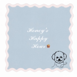 May include: A light blue square cloth with a scalloped pink border. The text "Honey's Happy Hour" is embroidered in white script. A small illustration of a dog and a cupcake are also featured.