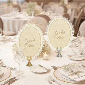 May include: Elegant table setting with two oval table number cards. The cards have a cream-colored background with the words "Table One" and "Table Two" in gold script. Each card is framed with a delicate lace border. The setting includes glassware, plates, and silverware.
