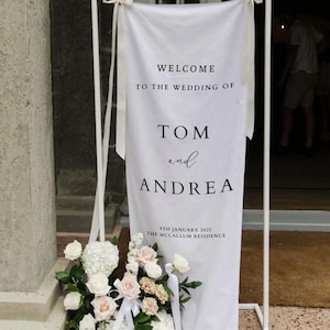 Op de afbeelding: Witte bruiloft welkom bord met zwarte tekst. Het bord staat "Welkom op de bruiloft van Tom en Andrea, 8 januari 2025, The McCallum Residence".