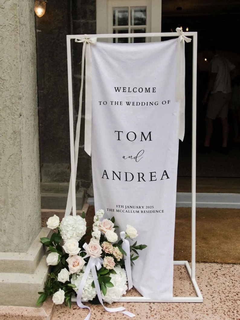 Op de afbeelding: Witte bruiloft welkom bord met zwarte tekst. Het bord staat "Welkom op de bruiloft van Tom en Andrea, 8 januari 2025, The McCallum Residence".