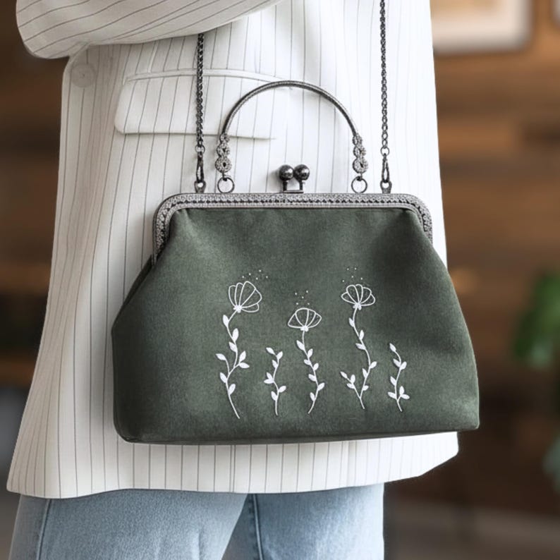 May include: A dark green handbag with a metal clasp and chain strap. The bag has a white floral embroidery design. The handle is a curved metal bar. The bag is held by a person wearing a white blazer.