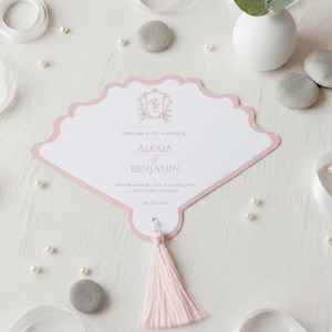 Può includere: Un cartoncino per il programma di matrimonio a forma di ventaglio con un bordo rosa chiaro e una nappa rosa chiaro abbinata. Il cartoncino bianco presenta uno stemma decorativo, il testo Welcome to the wedding of ALEXIA & BENJAMIN e la data 09.30.2023.