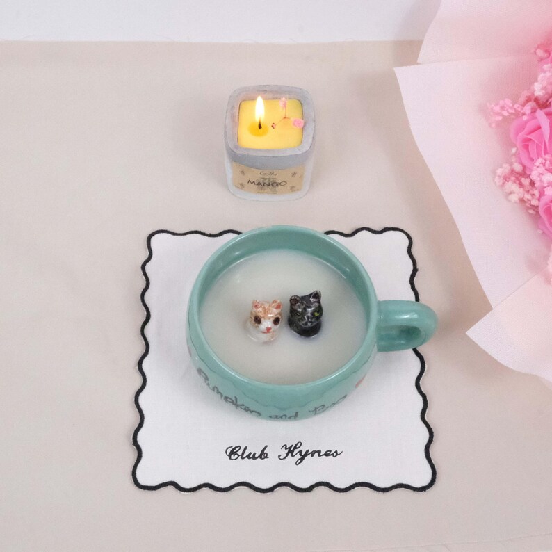 May include: A white ceramic mug with a black and white cat figurine inside. The mug is sitting on a white coaster with black trim and the text "Club Hynes" written on it. A small yellow candle with the word "MANGO" on it is in the background.