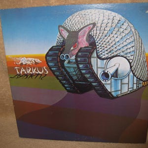 Pu&ograve; includere: Copertina di un album con un'immagine surreale di una creatura simile a un armadillo con cingoli. La creatura &egrave; ambientata su uno sfondo con un cielo blu e un primo piano geometrico colorato. Il titolo dell'album "Tarkus" &egrave; visibile.