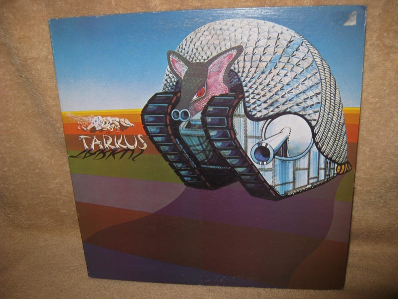 Pu&ograve; includere: Copertina di un album con un'immagine surreale di una creatura simile a un armadillo con cingoli. La creatura &egrave; ambientata su uno sfondo con un cielo blu e un primo piano geometrico colorato. Il titolo dell'album "Tarkus" &egrave; visibile.