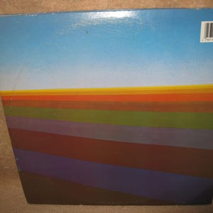 Pu&ograve; includere: Una copertina di un album vintage con un design sfumato. La parte superiore &egrave; un cielo azzurro chiaro, che sfuma in un orizzonte di strisce arancioni, gialle e verdi. Sotto, strisce angolate in tonalit&agrave; marroni, viola e blu creano un effetto a strati. Un codice a barre &egrave; visibile.