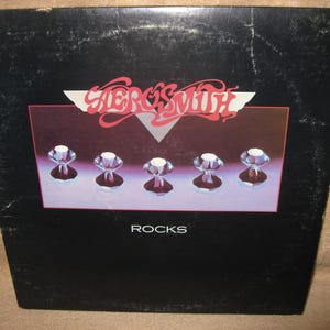 May include: Aerosmith's "Rocks" album cover. The album cover is black with a rectangular image in the center. The image features five diamond-shaped gems on a purple background. The band's name is in red and white script above the gems, with the word "ROCKS" in white below.