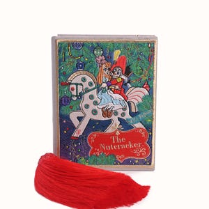 Könnte beinhalten: Eine dekorative Box mit einer farbenfrohen Illustration von Figuren aus "Der Nussknacker" auf einem Pferd, mit einer roten Quaste. Die Box hat ein festliches Design mit Weihnachtselementen und dem Titel "The Nutcracker".