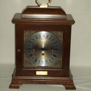 May include: A dark brown wooden mantel clock with a gold-colored handle. The clock face has Roman numerals and decorative gold accents. A small rectangular plaque is below the clock face.
