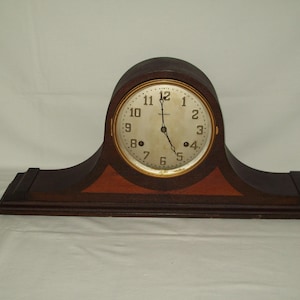 May include: A dark brown wooden mantel clock with a curved top and a rectangular base. The clock face is round with a gold-colored rim, featuring black numerals and hands. The clock is set to approximately 5:00.