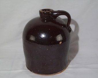 May include: A brown glazed stoneware jug with a handle. The jug has a rounded body and a slightly flared lip.