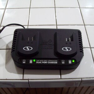 Może przedstawiać: Czarna ładowarka dwuportowa z dwoma portami ładowania. Ładowarka ma czarny przewód i napis "24V MAX ION + LITHIUM ION DUAL PORT CHARGER". Ładowarka ma dwie zielone kontrolki.