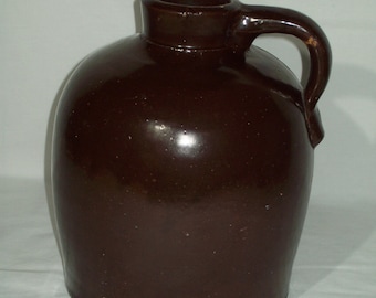 May include: A brown glazed stoneware jug with a handle. The jug has a rounded body and a wide mouth.