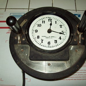 May include: A black and silver time clock with two levers. The clock face is white with black numbers and hands. The clock face reads "Gorrell & Gorrell, Hamirth, N.J."