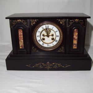 May include: A black marble mantel clock with gold accents and a white face. The clock has Roman numeral hour markers and a decorative design on the front.