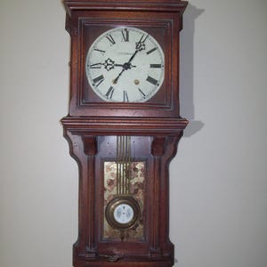 May include: A vintage wooden wall clock with a pendulum and Roman numeral face. The clock has a brass colored trim and a gold colored background behind the pendulum.