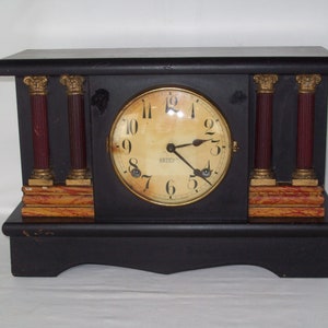 May include: A black wooden mantel clock with gold accents and a white face. The clock has Roman numerals and a gold-colored pendulum. The clock is sitting on a white surface.