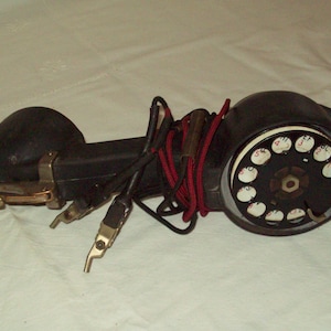 May include: An antique black telephone with a rotary dial. The phone has a receiver, a cradle, and a dial with white numbers. The phone's cord is wrapped with a red and black cord. The phone is resting on a white surface.