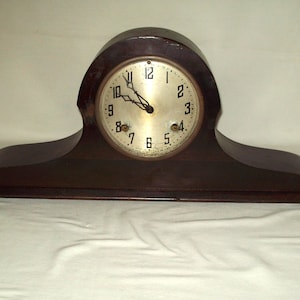 May include: A dark brown wooden mantel clock with a round clock face. The clock face has black numerals and hands. The clock has a curved top and a rectangular base. The clock is approximately 18 inches wide.