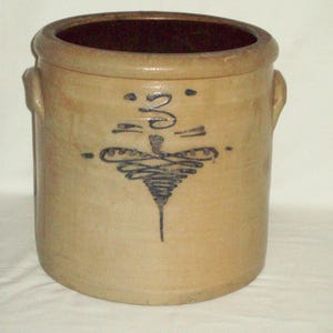 May include: Tan stoneware crock with a dark brown interior and two side handles. The crock features a blue decorative design on the front. The design appears to be a stylized floral or abstract motif. The crock is cylindrical in shape.
