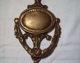 May include: A vintage brass door knocker with an ornate design. The knocker features a circular plate with a raised border and a decorative handle.