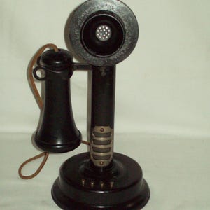 Puede incluir: Un teléfono antiguo negro de tipo vela con un auricular redondo, una boquilla y una base. El teléfono tiene un mango curvo y un cable. El auricular tiene el texto "Kellogg Switchboard & Supply Company".