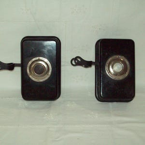 Pode incluir: Dois objetos retangulares pretos com um centro circular prateado e preto e uma alça preta. Os objetos são colocados lado a lado sobre uma superfície branca. Os objetos parecem ser vintage.