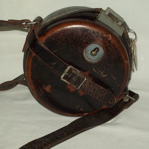 May include: A vintage, round leather case with a metal top and keyhole. The case is dark brown with a leather strap and buckle. A metal clasp and keys are attached to the side.