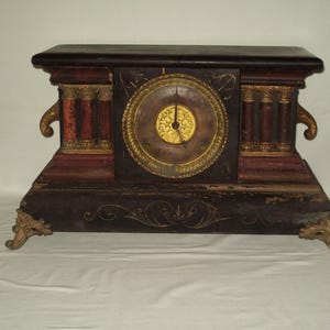 May include: Antique mantel clock with a dark wood finish, featuring a round clock face with gold accents. The clock has decorative columns and ornate gold-colored feet. The clock's face has gold hands and a gold-colored center.