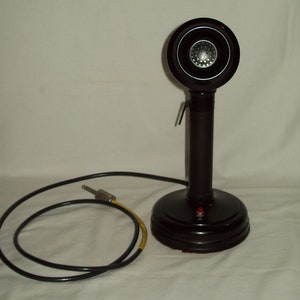 Puede incluir: Un micrófono vintage negro con una cabeza redonda y un cable negro. El micrófono tiene un botón rojo en la base.