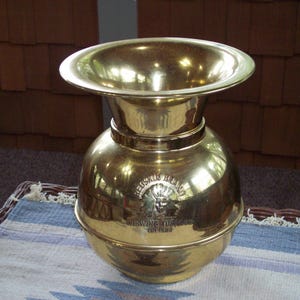 Puede incluir: Un recipiente de latón vintage con una parte superior ancha y acampanada y un cuerpo redondeado. El recipiente tiene una superficie brillante y reflectante y presenta el texto "REUSKIN BRAND" y "CHEWING TOBACCO" con un logotipo de sol. Es probable que sea un tarro de tabaco.