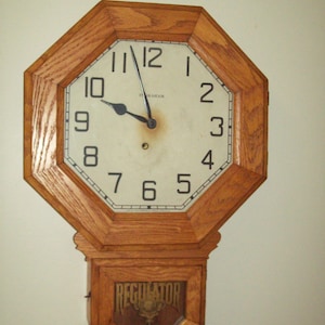 May include: An octagonal wooden wall clock with a light-colored face and black numerals. The clock has a rectangular lower section with the word "REGULATOR" visible. The clock hands point to approximately 9:58.